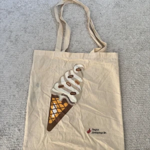 Beige tygkasse med glassmotiv - Snygg beige shoppingväska i bomull med ett stort tryck av en mjukglass på framsidan. Väskan har långa handtag och är perfekt att bära till skolan, stan eller stranden. Tryck med texten 'Region Jönköpings län' i hörnet.