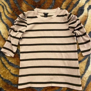 Randig blustopp med puffärm - Ljuslila blustopp med svarta horisontella ränder och trendiga puffärmar. Modellen har trekvartsärm och en rak passform. Perfekt för dig som gillar en stilren men ändå cool look.