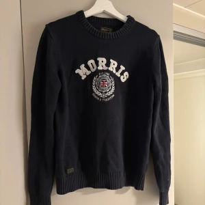 Mörkblå stickad tröja från Morris - Mörkblå stickad tröja från Morris med broderad logga och emblem framtill. Tröjan har rund halsringning, ribbade muddar och lång ärm.