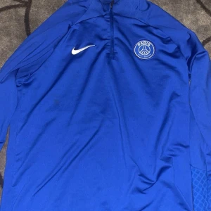 Blå PSG långärmad träningströja Nike - Säljer en blå långärmad fotbollströja från Nike med PSG-logga på bröstet. Tröjan har halv dragkedja, Dri-FIT-material och slim fit-passform. Perfekt för träning eller när du vill representera Paris Saint-Germain.