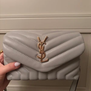 Grå YSL axelväska  - Supersnygg grå axelväska från YSL i quiltat skinn med guldiga detaljer och ikonisk YSL-logga framtill. Väskan har justerbar axelrem och praktiskt innerfack med dragkedja. Perfekt för dig som vill ha en stilren och lyxig accessoar.