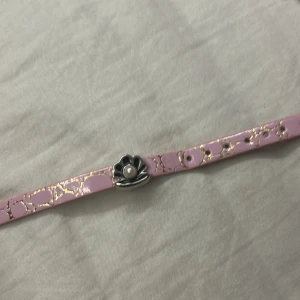 Rosa armband med blomma och pärla - Sött armband i ljusrosa med guldiga detaljer och ett silverfärgat blomspänne med en liten pärla i mitten. Armbandet har ett unikt mönster och passar perfekt för dig som gillar gulliga accessoarer med en touch av elegans.