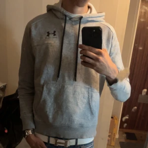 Grå hoodie från Under Armour - Säljer en grå hoodie från Under Armour med svart logga på bröstet. Tröjan har huva med svarta snören, magficka och långa ärmar. Perfekt för chill eller träning, riktigt skön och stilren modell. Unik underarmour modell så säljer lite dyrare