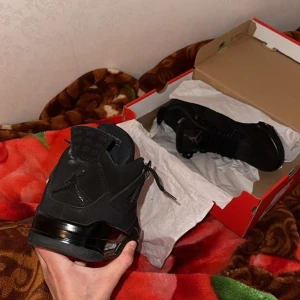 Svarta Nike Air Jordan 4 sneakers - Säljer ett par helsvarta Nike Air Jordan 4 sneakers med klassisk siluett och Jumpman-logga på hälen. Skorna har snörning, meshpaneler och detaljer i mocka och syntet. Perfekta för dig som vill ha en clean och stilren look med ikonisk streetwear-vibe.
