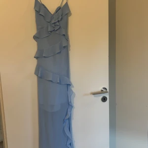 Ljusblå långklänning från Vero Moda - Supersnygg ljusblå långklänning från Vero Moda med volangdetaljer och smala axelband. Klänningen har omlottkänsla upptill, snörning i ryggen och är tillverkad i ett lätt, flowy material som ger en drömmig look. Perfekt för dig som vill sticka ut med en romantisk vibe.