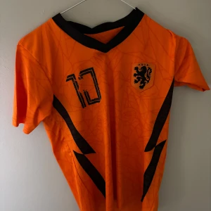 Holland Memphis Depay fotbollströja - Snygg orange Holland fotbollströja med svarta detaljer, nummer 10 och Memphis på ryggen. KNVB-logga på bröstet och coola svarta mönster längs sidorna. Perfekt för dig som älskar fotboll och vill sticka ut på planen eller läktaren.