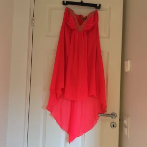 Rosa festklänning från Lipsy London - Supersnygg rosa klänning från Lipsy London med assymetrisk fåll och v-ringning dekorerad med glittriga pärlor. Klänningen är axelbandslös och har ett lätt, flowy tyg som ger en riktigt cool look. Perfekt för dig som vill sticka ut!