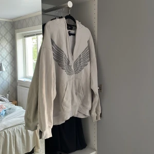 Grå hoodie med vingar och dragkedja - Säljer en oversized ljusgrå hoodie från H&M med stort tryck av vingar på bröstet. Tröjan har huva med snören, dragkedja framtill och en stor känguruficka. Perfekt för chill dagar eller när du vill sticka ut lite extra. 🫶