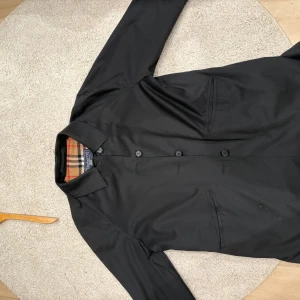 Svart Burberrys rock  - Klassisk svart rock från Burberrys med ikoniskt rutig foder i beige, svart, vitt och rött. Yttertyg i polyester och bomull, fodret är 100% ull. Rak passform, knappar framtill och två fickor. Perfekt för dig som gillar tidlös brittisk stil.