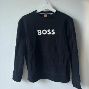 Svart sweatshirt från BOSS - Svart sweatshirt från BOSS med vit BOSS-logga framtill. Klassisk rund halsringning och långa ärmar. Tillverkad i mjuk bomullsmix som är skön att bära. Perfekt för en chill och stilren look. Storlek S