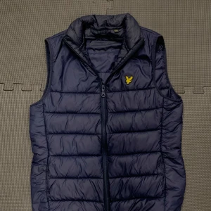 Mörkblå dunväst från Lyle & Scott XS - Snygg mörkblå dunväst från Lyle & Scott med gul logga på bröstet. Västen har quiltad design, hög krage och dragkedja framtill. Perfekt för lager-på-lager och ger en sportig vibe till din outfit.