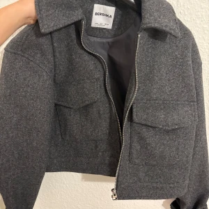 Grå croppad jacka från Bershka - Snygg grå croppad jacka från Bershka med klassisk krage, två stora bröstfickor och dragkedja framtill. Jackan har en loose fit och är tillverkad i ullblandning, perfekt för höst och vinter. Enkel och stilren design som lyfter vilken outfit som helst.