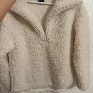 Beige teddytröja från Gina Tricot - Supermjuk och fluffig beige teddytröja från Gina Tricot med halv dragkedja och hög krage. Perfekt för kyliga dagar, har lång ärm och loose fit. Materialet är fleece som ger en mysig känsla.