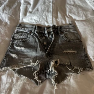 Gråa slitna highwaist jeansshorts - Säljer ett par gråa högmidjade jeansshorts med råa kanter och slitningar framtill. Shortsen har knappar i gylfen, klassiska fickor och en cool, edgy look med mycket fransar och trasiga detaljer. Perfekta för sommaren och festivaler.