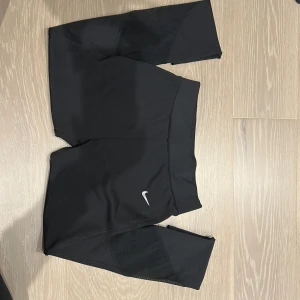 Svarta träningstights-shorts från Nike - Svarta, tighta träningstights-shorts från Nike med klassisk vit swoosh-logga på ena benet. De har hög midja och är gjorda i ett stretchigt syntetmaterial som passar perfekt för träning. Snygg och sportig look.