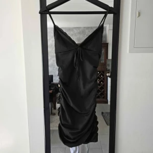 Svart satinklänning med snörning - Supersnygg svart klänning i satin med smala axelband och v-ringning. Klänningen har snörning framtill och draperad effekt längs sidorna som ger en cool och trendig look. Perfekt för dig som gillar stilrena och eleganta plagg.