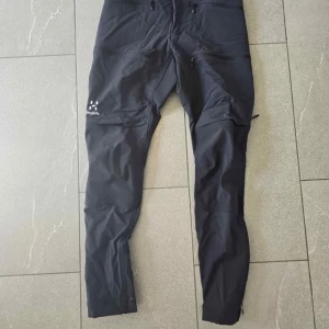 Svarta friluftsbyxor från Haglöfs dam - Svarta friluftsbyxor från Haglöfs i dammodell med flera dragkedjeförsedda fickor och smal passform. Byxorna är tillverkade i slitstarkt syntetmaterial och har diskret logga på låret. Perfekta för utomhusaktiviteter och äventyr. Använda 1 gång vid regn 