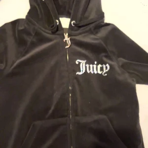 Juicy Couture svart velour hoodie XS - Svart hoodie i mjuk velour från Juicy Couture med glittrande stenar på huvan och texten 'Juicy' framtill. Dragkedja med J-berlock och kängurufickor. Perfekt för dig som vill ha den ikoniska 2000-talslooken.