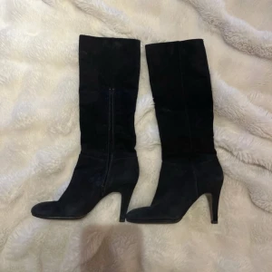 Svarta mockastövlar med klack  - Snygga svarta stövlar från indiska i mjuk mocka med hög klack och rund tå. Stövlarna har dragkedja på insidan och en stilren, klassisk look som passar till många outfits. Perfekta för dig som vill ha en elegant siluett och gillar boots med lite höjd.