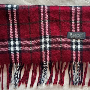 Rutig vinröd halsduk från Burberrys - Klassisk halsduk från Burberrys i vinrött, svart och vitt rutmönster. Gjord i 100% lammull med fransar i ändarna. Perfekt accessoar för att lyfta din vinterstil med en tidlös brittisk touch.