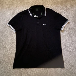 Hugo Boss Piké - Säljer en svart pikétröja från BOSS med vit logga broderad på bröstet. Pikén är i bra skick och passar storlek S-M. Pikén är köpt på Johnells i juni 2025. Perfekt för en clean och stilren look (Pris går att diskutera).