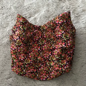 Blommig korsettopp från Zara - Säljer en färgstark korsettopp från Zara med blommigt mönster i rosa, grönt och rött på svart botten. Toppen är axelbandslös och har en snygg, figurnära passform. Perfekt för dig som vill sticka ut med en cool och trendig look.