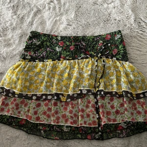 Blommig volangkjol från MNG, strl 36 - Supersöt kort volangkjol från MNG med flera lager och blommigt mönster i svart, gult, rött och grönt. Kjolen har knytband i midjan och är perfekt för dig som gillar färg och mönster. Lätt och luftig känsla, passar till sneakers eller sandaler.