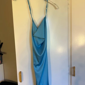 Blå långklänning från NAANAA - Säljer en snygg blå långklänning från NAANAA med tunna axelband och draperad framsida. Klänningen har öppen rygg med snörning och är gjord i ett glansigt satinliknande material. Perfekt för dig som vill sticka ut med en cool och trendig look.