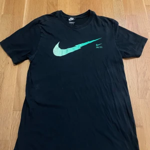 Svart Nike t-shirt med grön logga - Svart t-shirt från Nike i standard fit, med en stor grön blixtformad Swoosh-logga och trycket 20/21 på bröstet. Klassisk rund hals och korta ärmar, tillverkad i mjuk bomull. Perfekt för dig som gillar sportig och clean stil. Shorts kommer med.