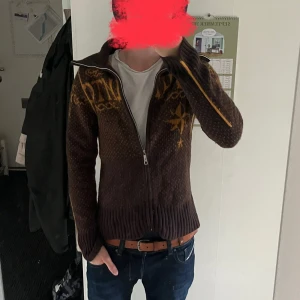 Unik Vintage Diesel tjocktröja - Säljer en mörkbrun stickad kofta från Diesel. Koftan har hel dragkedja, hög krage och ribbade muddar. Snygga detaljer längs ärmarna och coolt mönster på både fram- och baksida. Perfekt för dig som gillar unik stil! 100% wool. OBS!: Har krympt i tvätten är mer av en S/M (inte XL allstå)