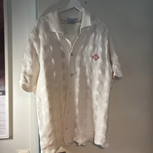 Vit stickad skjorta från Casablanca - Säljer en vit stickad skjorta från Casablanca med öppen stickning och vågigt mönster. Skjortan har korta ärmar, klassisk krage, knappar framtill och röd broderad logga på bröstet. Perfekt för dig som vill sticka ut med en unik och luftig look.