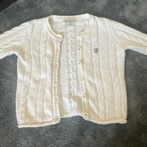 Vit kabelstickad kofta Hampton Republic - Vit kabelstickad kofta från Hampton Republic med rund halsringning och knappar framtill. Klassisk design med diskret HR-brodyr på bröstet. Perfekt att slänga över en t-shirt eller klänning för en clean look.