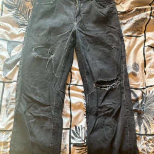 Svarta slitna jeans med hål på knäna - Säljer ett par svarta jeans med raka ben och slitna detaljer på båda knäna. Jeansen har klassisk femficksdesign, dragkedja och knapp framtill. Perfekta för en avslappnad och edgy stil. Lågmidjade.