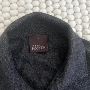 Mörkgrå merinoullskofta Oscar Jacobson - Snygg mörkgrå kofta från Oscar Jacobson i 100% merinoull. Klassisk modell med krage, knäppning framtill och två fickor. Perfekt för lager-på-lager och riktigt skön att ha på sig under kyliga dagar.