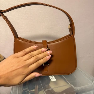 Brun axelväska i skinn med gulddetaljer - Snygg brun axelväska i skinn med stilrena gulddetaljer och justerbart axelband. Väskan har en minimalistisk design med rundade hörn och en praktisk dragkedja upptill. Perfekt storlek för det viktigaste och passar till många olika outfits.