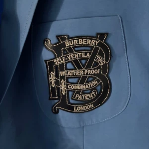Blå Burberry kavaj med emblem - Snygg blå kavaj från Burberry med stort broderat emblem på bröstfickan och klassiskt rutigt foder på insidan. Kavajen har två knappar framtill och raka fickor. Perfekt för dig som vill sticka ut med en exklusiv och stilren look.
