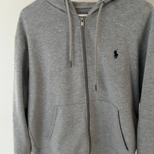 Grå hoodie från Polo Ralph Lauren - Grå hoodie från Polo Ralph Lauren med dragkedja, snörning i huvan och klassisk svart logga på bröstet. Tröjan har två fickor framtill och är tillverkad i mjuk bomullsmix. Perfekt för chill dagar och enkel att matcha med jeans eller joggers.