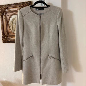 Beige kappa från Zara med dragkedja - Stilren beige kappa från Zara med diskret fiskbensmönster. Jackan har rund halsringning, hel dragkedja framtill och två snedställda fickor med dragkedjor. Perfekt för dig som gillar en clean look med snygga detaljer. Använd ett par gånger, nytt skick