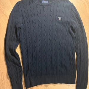 Mörkblå kabelstickad tröja från Gant - Kabelstickad mörkblå tröja från Gant i premium bomull. Klassisk rund halsringning, ribbade muddar och diskret broderad logga på bröstet. Perfekt för dig som gillar stilren och tidlös design.