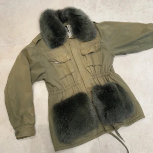 Olivgrön pälsjacka Windie Custommade - Säljer en så fin olivgrön jacka från Custommade med fluffig pälsdetalj vid krage och fickor. Jackan har knappar, dragsko i midjan och två bröstfickor med lock. Det har bara släppts 59 exemplar av denna jackan då den kommer från deras mer exklusiva serie. Skick enligt bilder, är ljusare på missa delar vilket man kan se i bilden. 
