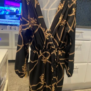 Svart omlottklänning med guldmönster - Svart kort omlottklänning med långa ärmar och snyggt guldigt kedjemönster. Djup v-ringning och knyt i midjan för en smickrande passform. Perfekt för dig som gillar statement-plagg med lyxig vibe.