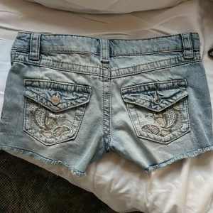 Ljusblå jeansshorts från Gina Tricot - Snygga ljusblå jeansshorts från Gina Tricot med slitna detaljer och råa kanter. Shortsen har fickor med broderi och knappar bak, samt klassisk femficksdesign och bälteshällor. Perfekta för en chill sommardag.