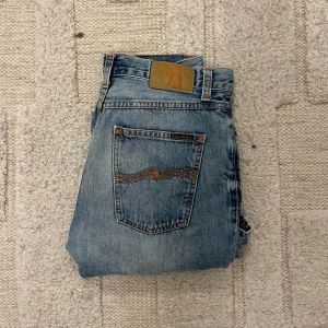 Ljusblå jeans från Nudie Jeans, 30/30 - Säljer ett par ljusblå jeans från Nudie Jeans med klassisk femficksdesign och orangea sömmar. Jeansen har knäppning med knappar och snyggt sliten tvätt. Perfekta för dig som gillar en avslappnad och tidlös look.