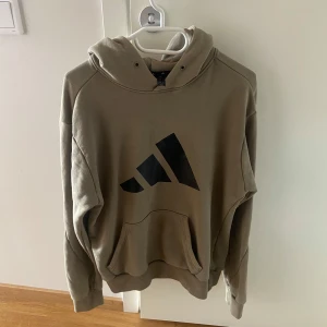Beige hoodie från Adidas med logga - Säljer en beige hoodie från Adidas med stor svart logga på bröstet. Tröjan har huva med snörhål, känguruficka och långa ärmar. Tillverkad i bomull och polyester, perfekt för chill dagar eller träning.