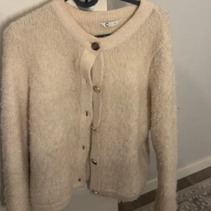 Beige fluffig kofta med knappar - Mysig beige kofta med fluffig känsla och stora knappar framtill. Rund halsringning och långärmad, stilren design som passar till det mesta.