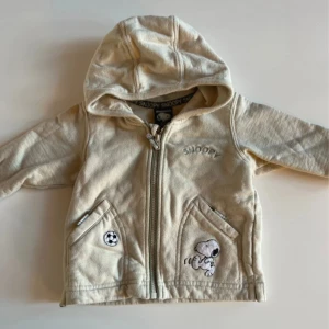 Beige Snoopy-hoodie från H&M - Supersöt beige hoodiejacka från H&M med broderad Snoopy och fotbollsmotiv både fram och bak. Jackan har huva, dragkedja och två fickor framtill. Perfekt för små Peanuts-fans som vill ha en mysig och lekfull stil.