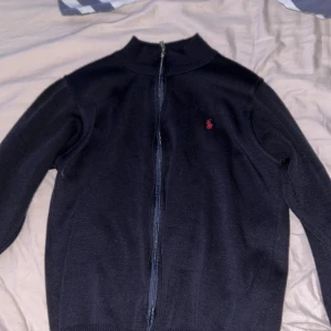 Polo Ralph Lauren fullzip hoodie - Mörkblå tröja från polo Ralph Lauren med hel dragkedja framtill och klassisk röd logga på bröstet. Tröjan har ribbade muddar och krage, långärmad och tillverkad i mjukt stickat material. Perfekt för dig som gillar stilren och enkel design.