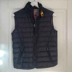 Svart dunväst från Parajumpers - Svart quiltad dunväst från Parajumpers med hög krage, dragkedja framtill och två snedställda fickor med dragkedja. Västen har en gul detalj vid halsen och är superlätt. Perfekt för lager-på-lager under kyliga dagar.