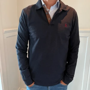 Mörkblå pikétröja från Polo Ralph Lauren - Snygg mörkblå långärmad pikétröja från Polo Ralph Lauren med röd broderad logga på bröstet. Slim fit-modell i stretchig bomullsmix, klassisk krage och knappar framtill. Perfekt för dig som gillar stilrena och sportiga plagg. Priset går att diskuteras vid snabb affär 