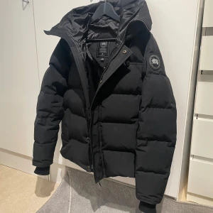 Svart pufferjacka från Canada Goose - Säljer en svart pufferjacka från Canada Goose med klassisk patch på ärmen. Jackan har huva, dragkedja och knappar framtill samt ribbade muddar vid ärmsluten. Perfekt för kalla dagar och riktigt snygg streetstil. Storleken på jackan är M som man kan se men jag skulle säga att den passar S mycket bra också
